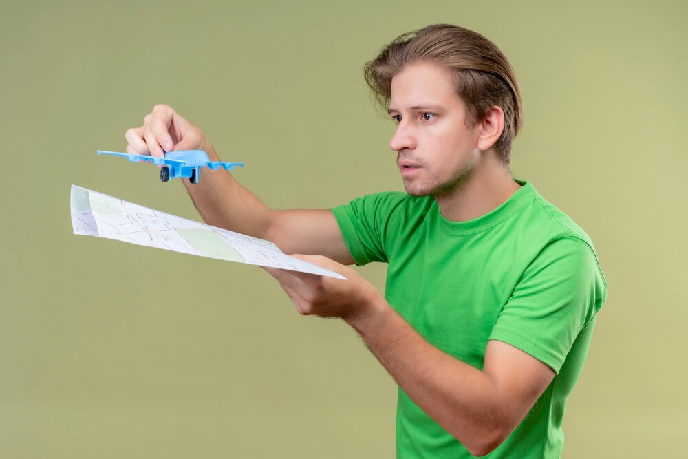Young handsome man wearing green t-shirt holding toy airplane and map with serious expression on face standing over green wall