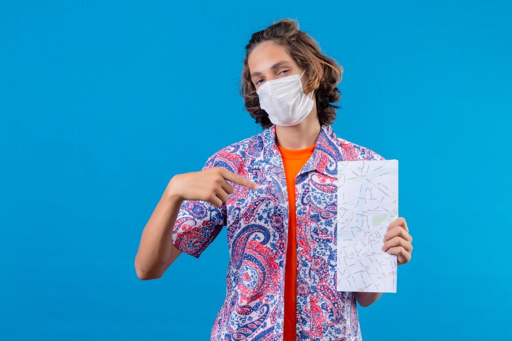 Young handsome guy wearing facial protective mask holding air tickets pointing fith finger to himself proud and confident smiling standing